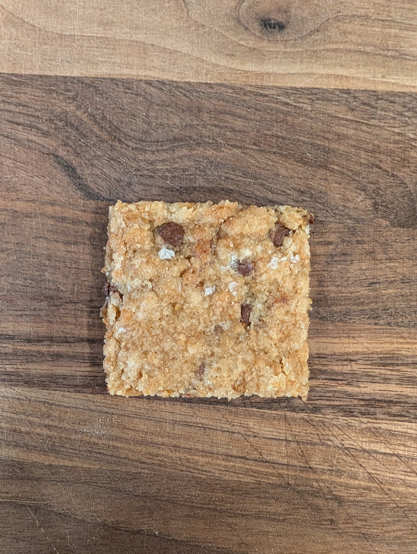 Single square piece of chocolate chip cookie brittle with flaky sea salt and visible chocolate chips on walnut cutting board, overhead view