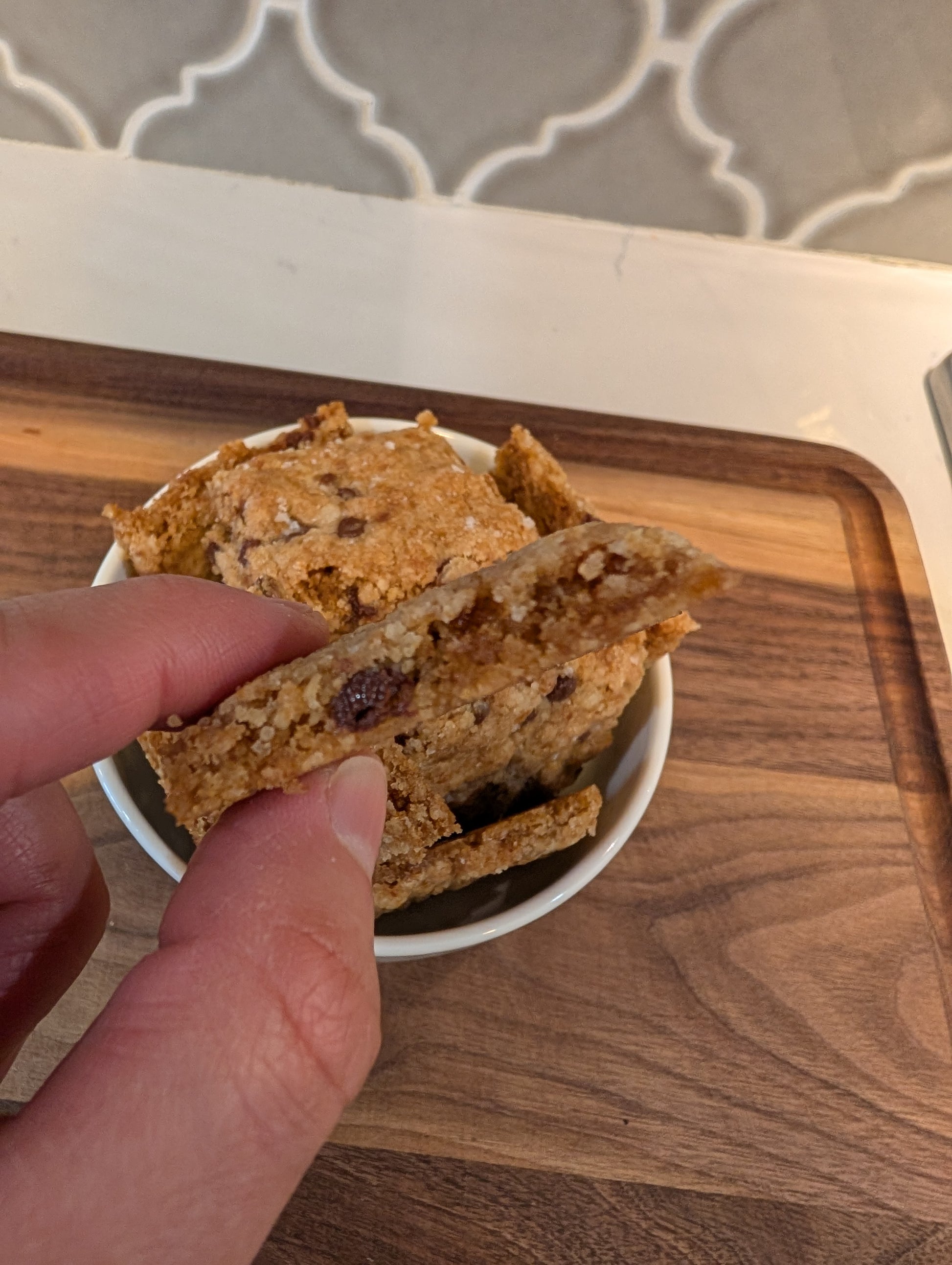 Hand holding a piece of chocolate chip cookie brittle showing thin crispy edge and embedded chocolate chips, with bowl of brittle pieces in background on walnut cutting board