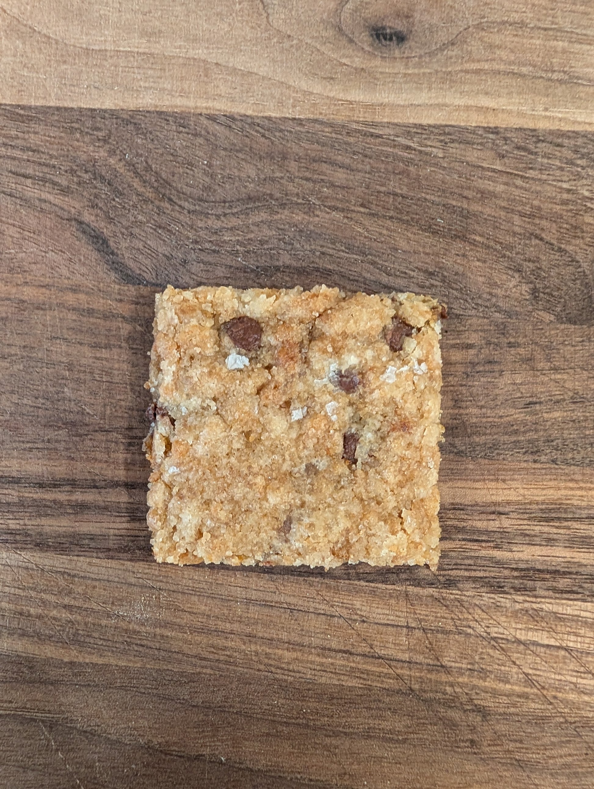 Single square piece of chocolate chip cookie brittle with flaky sea salt and visible chocolate chips on walnut cutting board, overhead view