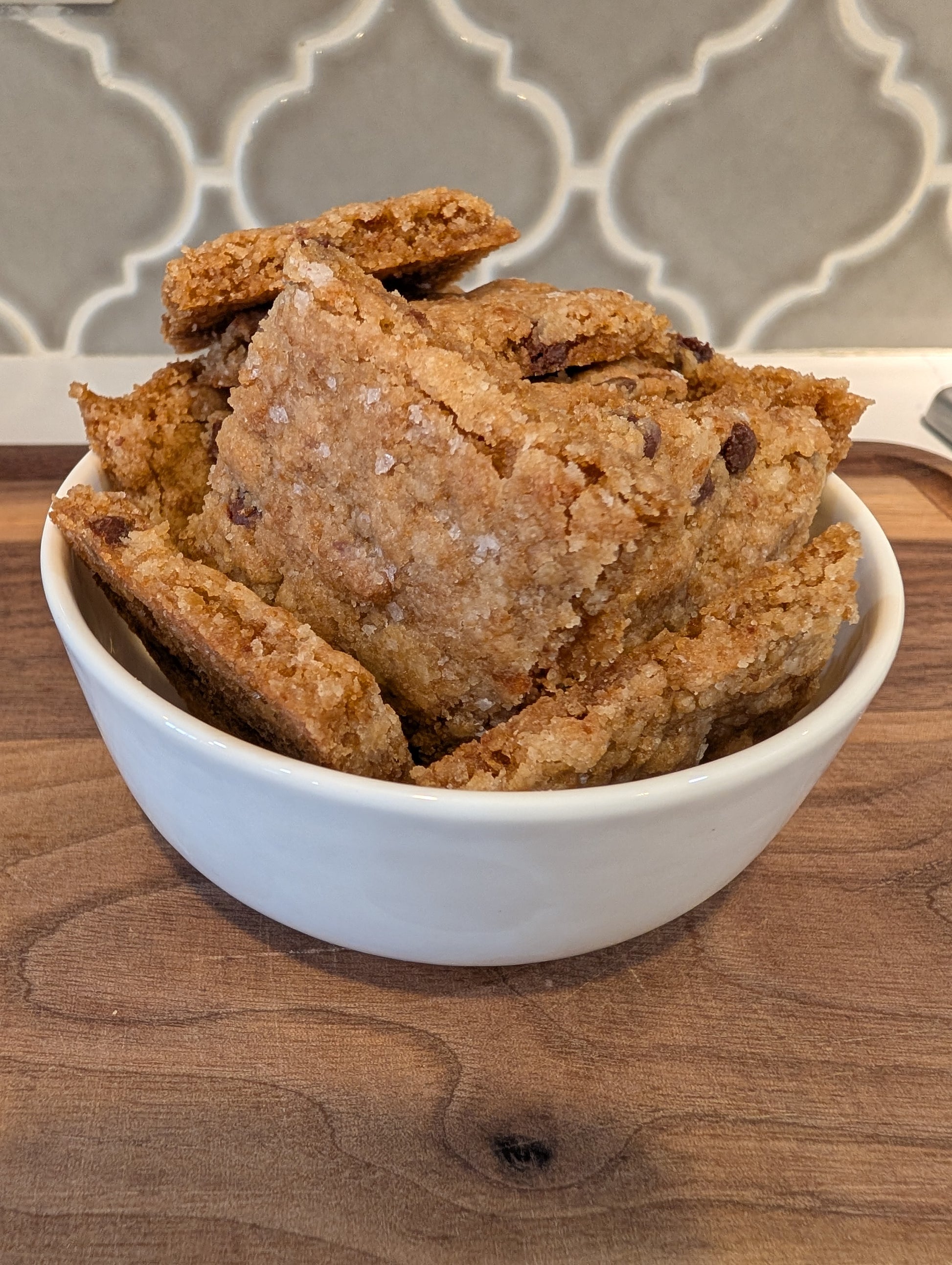 White bowl filled with golden-brown chocolate chip cookie brittle pieces topped with flaky sea salt, showing thin crispy texture with visible chocolate chips throughout